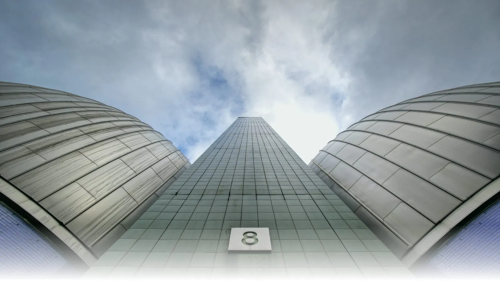 Looking up between two stainless-steel egg-shaped anaerobic digesters at the Newtown Creek Wastewater Treatment Plant in Brooklyn, New York, with the central tower labelled '8' rising into a cloudy sky