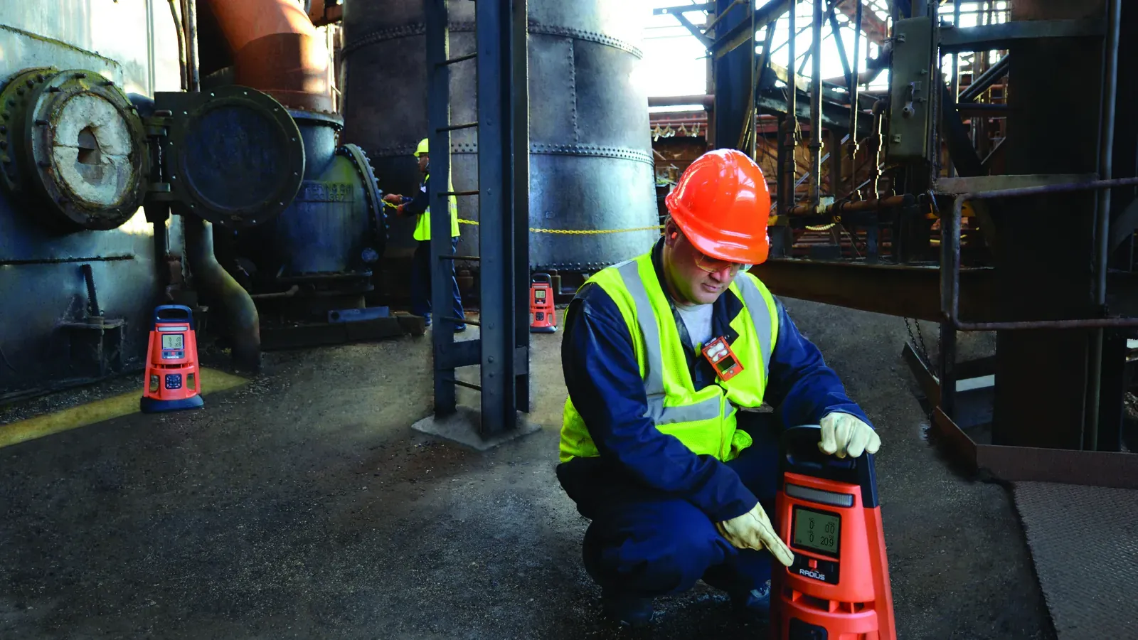Industrial Scientific Radius BZ1 area gas monitor deployed inside an active industrial plant by a technician in PPE, with two additional Radius units visible monitoring perimeter coverage