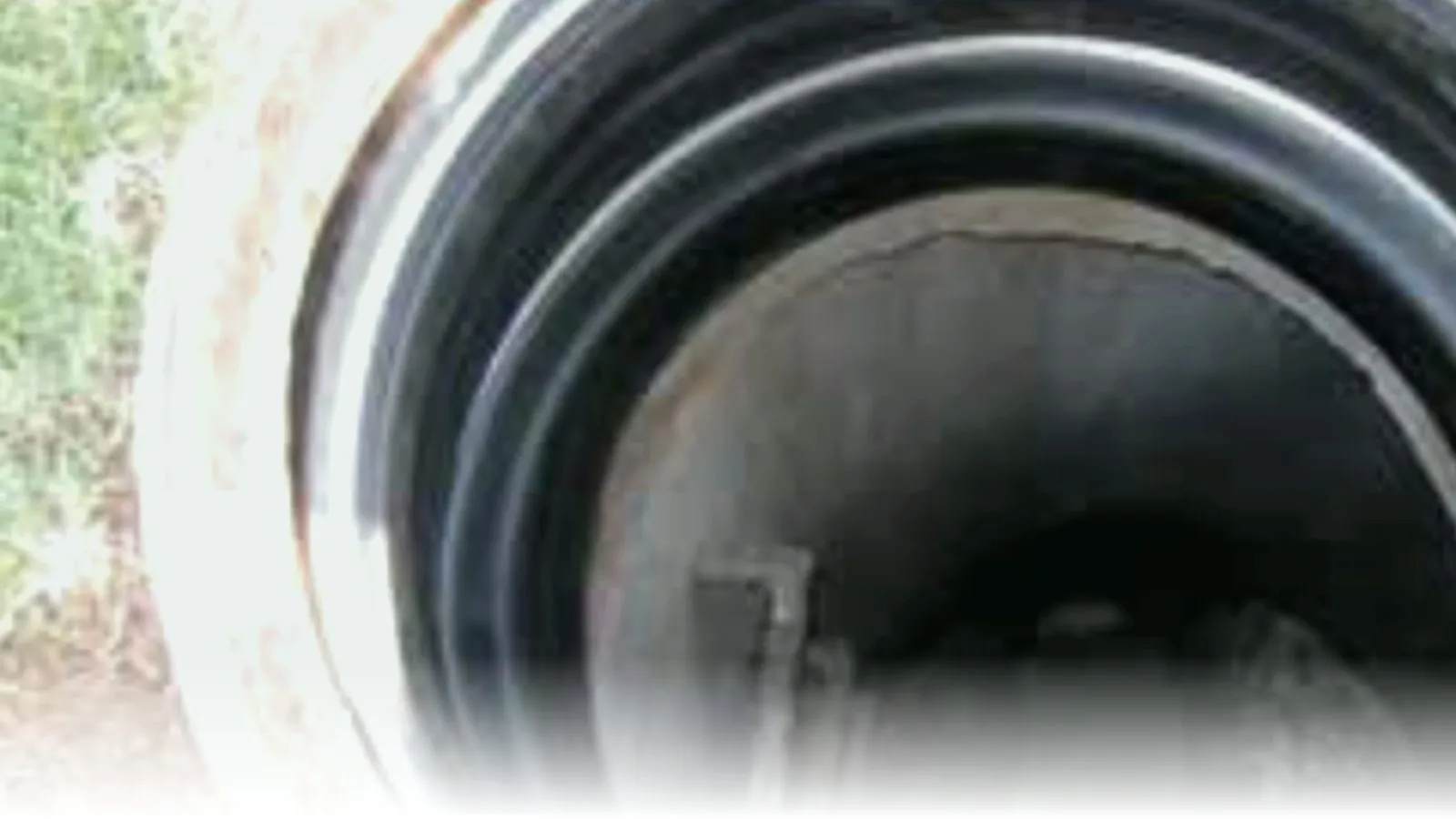 Cretex internal chimney seal installed inside a precast concrete manhole — black rubber seal extends from the cast-iron frame down into the chimney section, photographed from above with grass visible at the rim