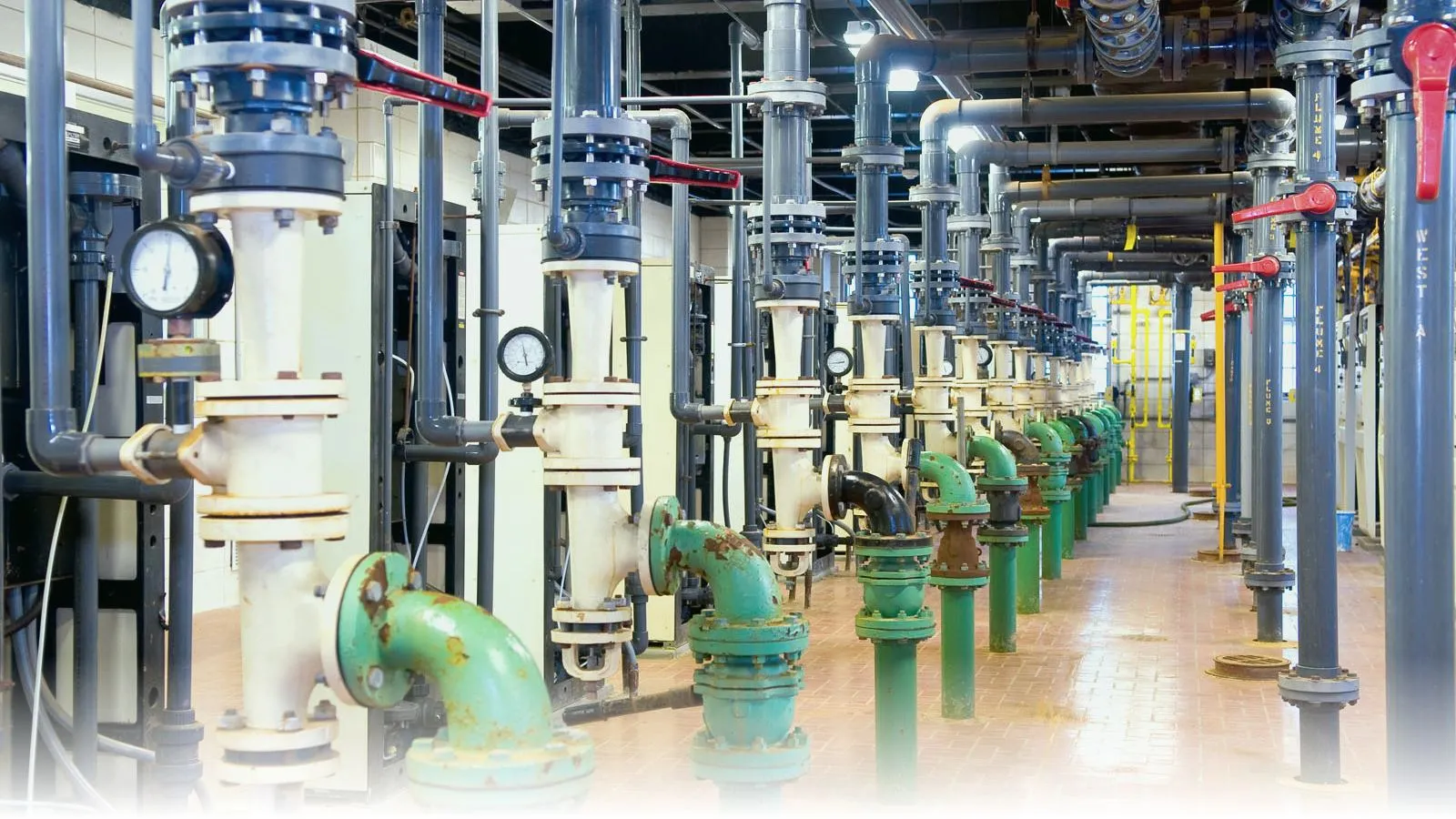Interior of a drinking-water treatment plant — rows of vertical filter columns with pressure gauges, green flanged pump piping, and overhead distribution mains on a brick floor