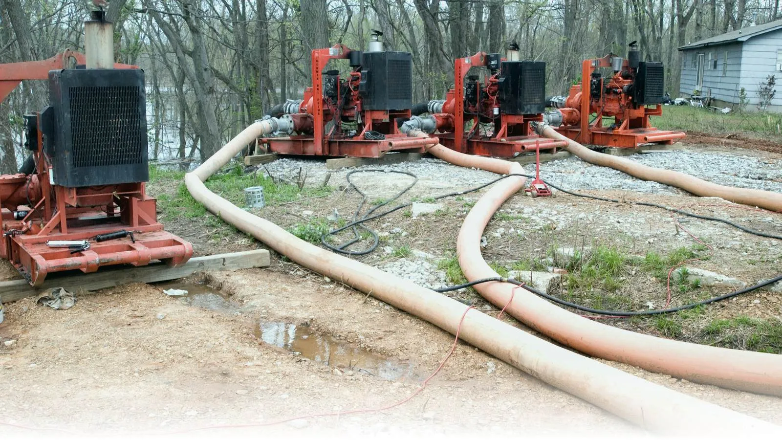 Four large orange diesel-engine emergency pumps lined up at a levee with flexible orange discharge hoses snaking across gravel during a Mississippi River flood response in Illinois