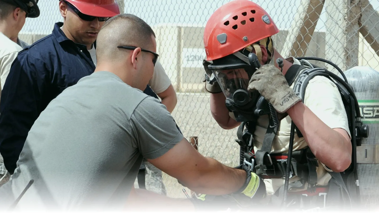 Rescue team prepping a worker for confined-space entry — the worker wears a red helmet, full-face SCBA mask, breathing apparatus tank, and harness while teammates check the rigging at the manhole